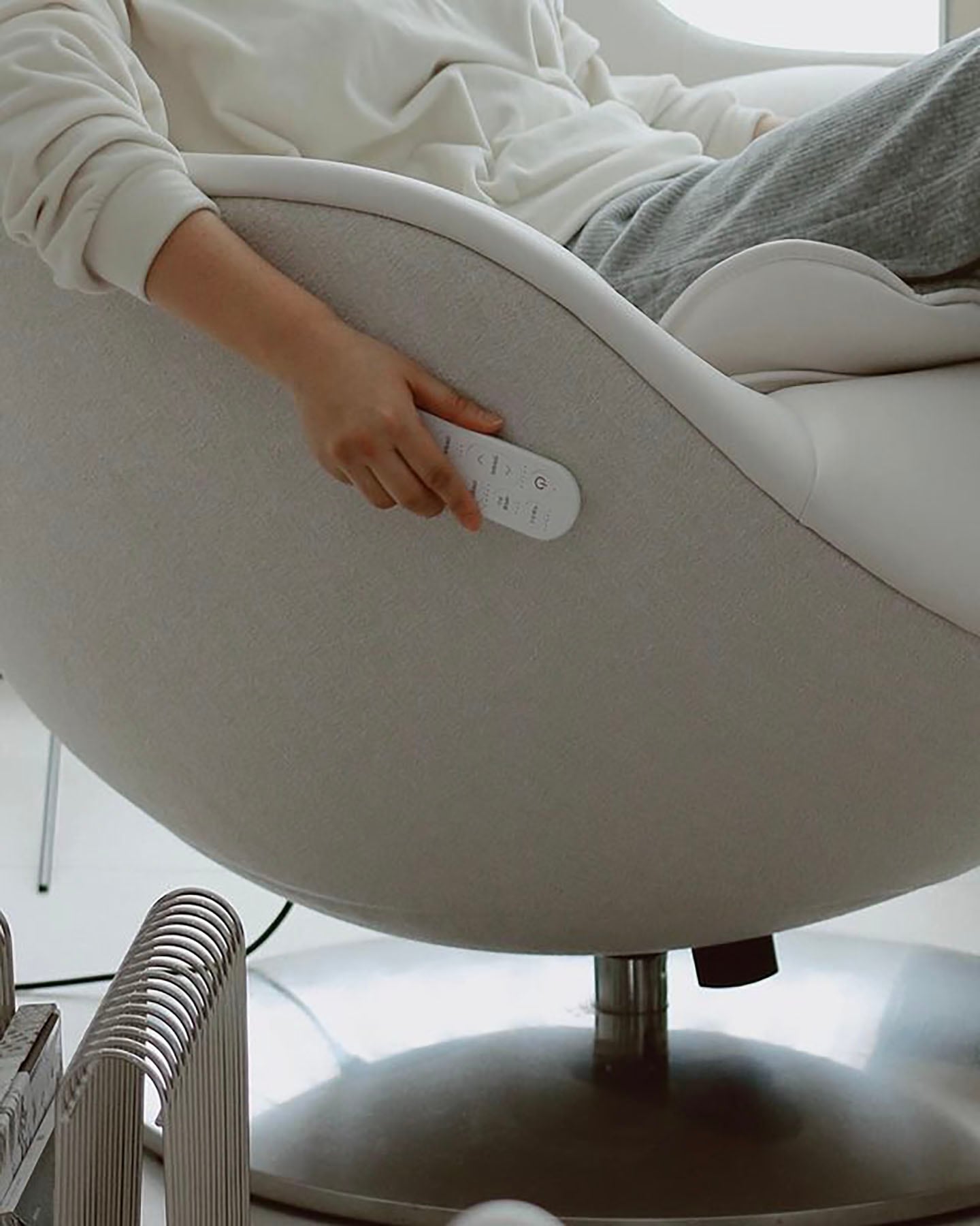 편안한 옷차림의 사람이 안마의자에 기대어 앉아 의자 옆면에 있는 리모컨을 손에 쥐고 조작하는 모습