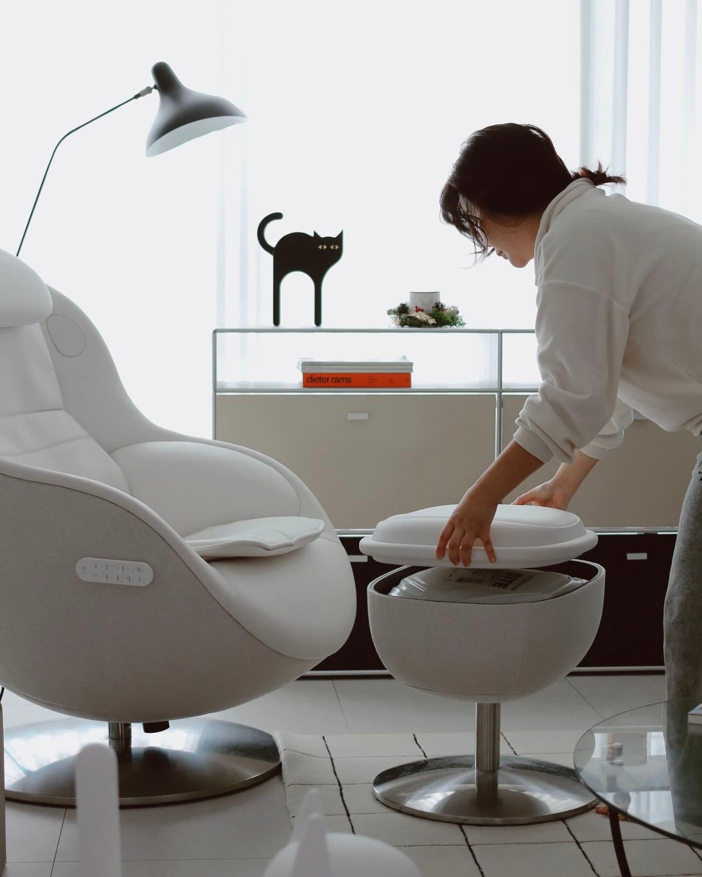 편안한 분위기의 거실에서 한 사람이 수납형 발받침대의 상단 쿠션을 들어 올려 내부 수납 공간을 확인하는 모습