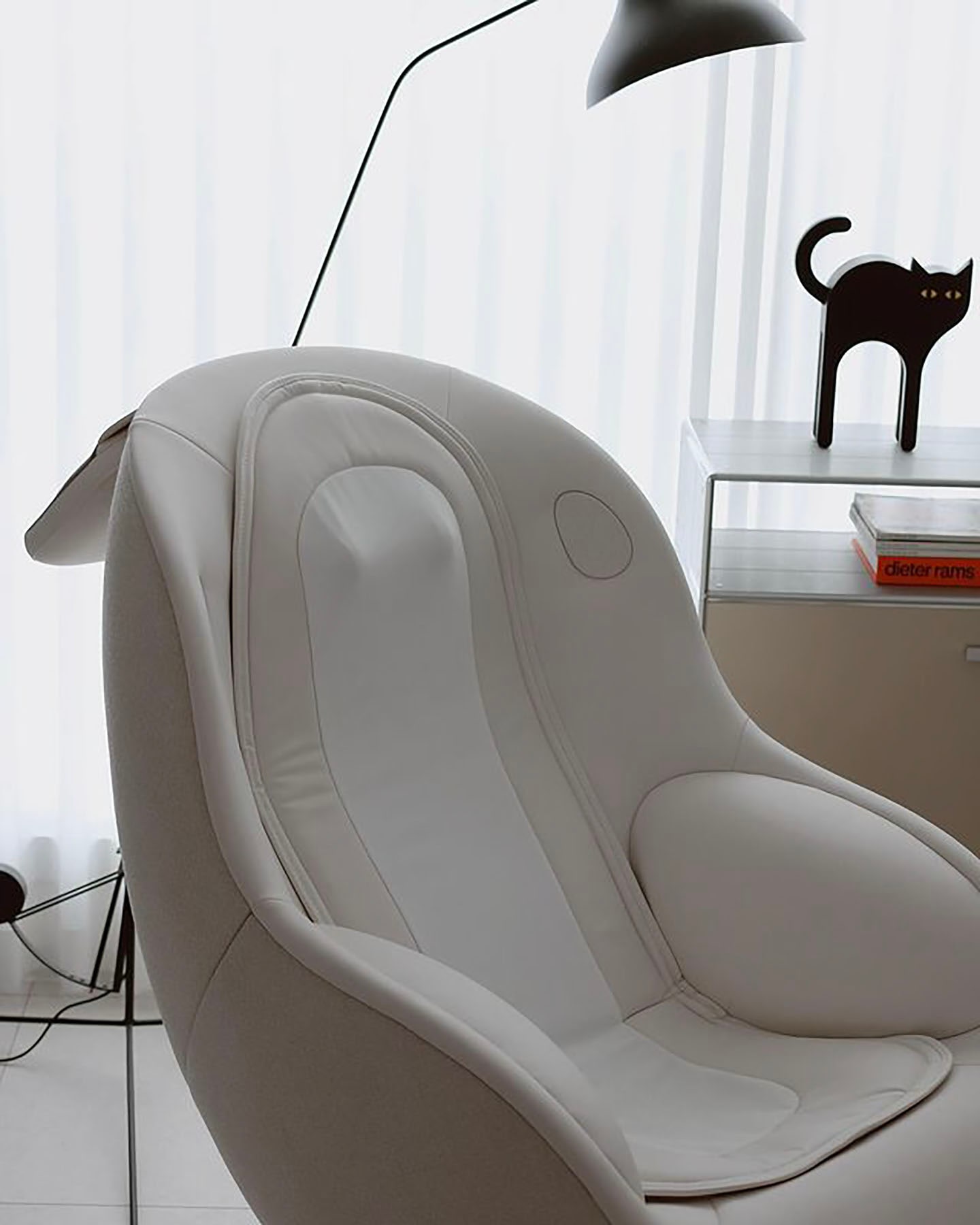 부드러운 곡선형 디자인의 안마 의자가 배치된 미니멀한 실내 공간으로, 옆에는 투명한 선반 위에 검은 고양이 장식품과 책이 놓여 있으며, 은은한 조명이 비추는 차분한 분위기의 이미지