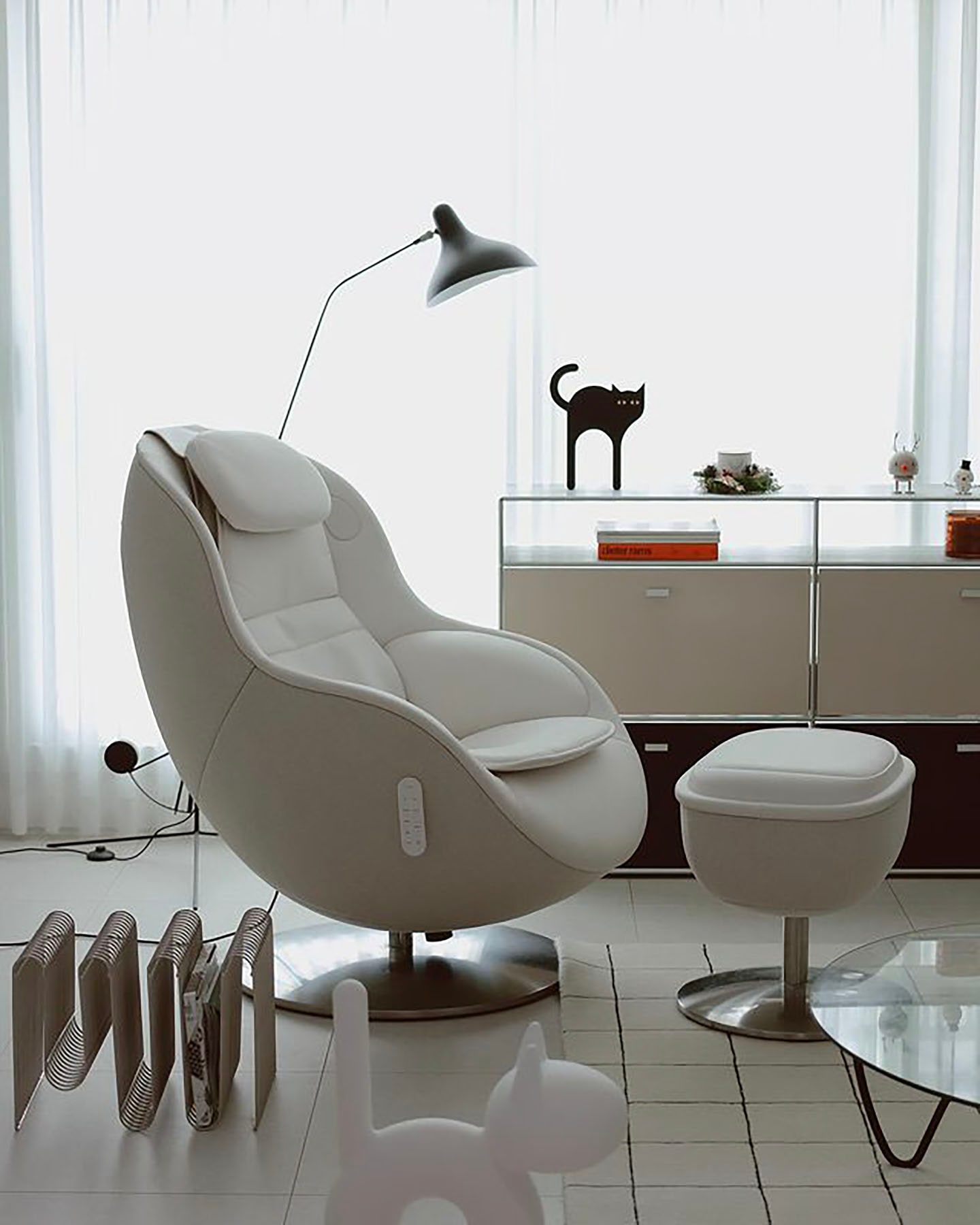 곡선형 디자인의 안마 의자와 오토만이 배치된 밝고 모던한 실내 공간으로, 옆에는 투명한 선반 위에 검은 고양이 장식품과 책이 놓여 있으며, 바닥에는 독특한 디자인의 책꽂이와 장식품이 배치된 미니멀한 분위기의 이미지