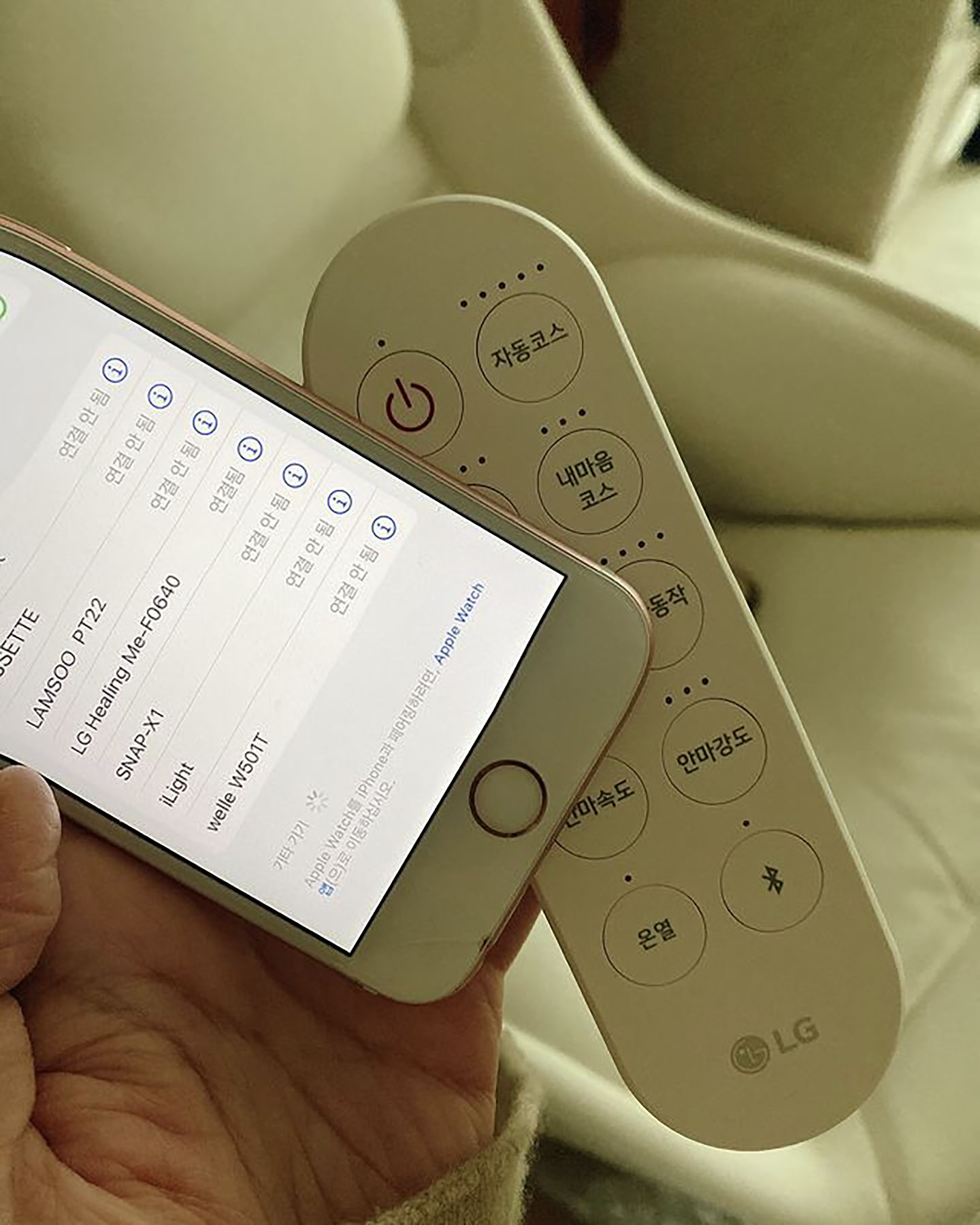 안마 의자의 무선 리모컨과 스마트폰을 함께 들고 있는 클로즈업 이미지로, 스마트폰 화면에는 블루투스 연결 목록이 표시되어 있으며, 리모컨에는 전원, 자동 코스, 안마 강도 조절 등의 버튼이 배열된 모습