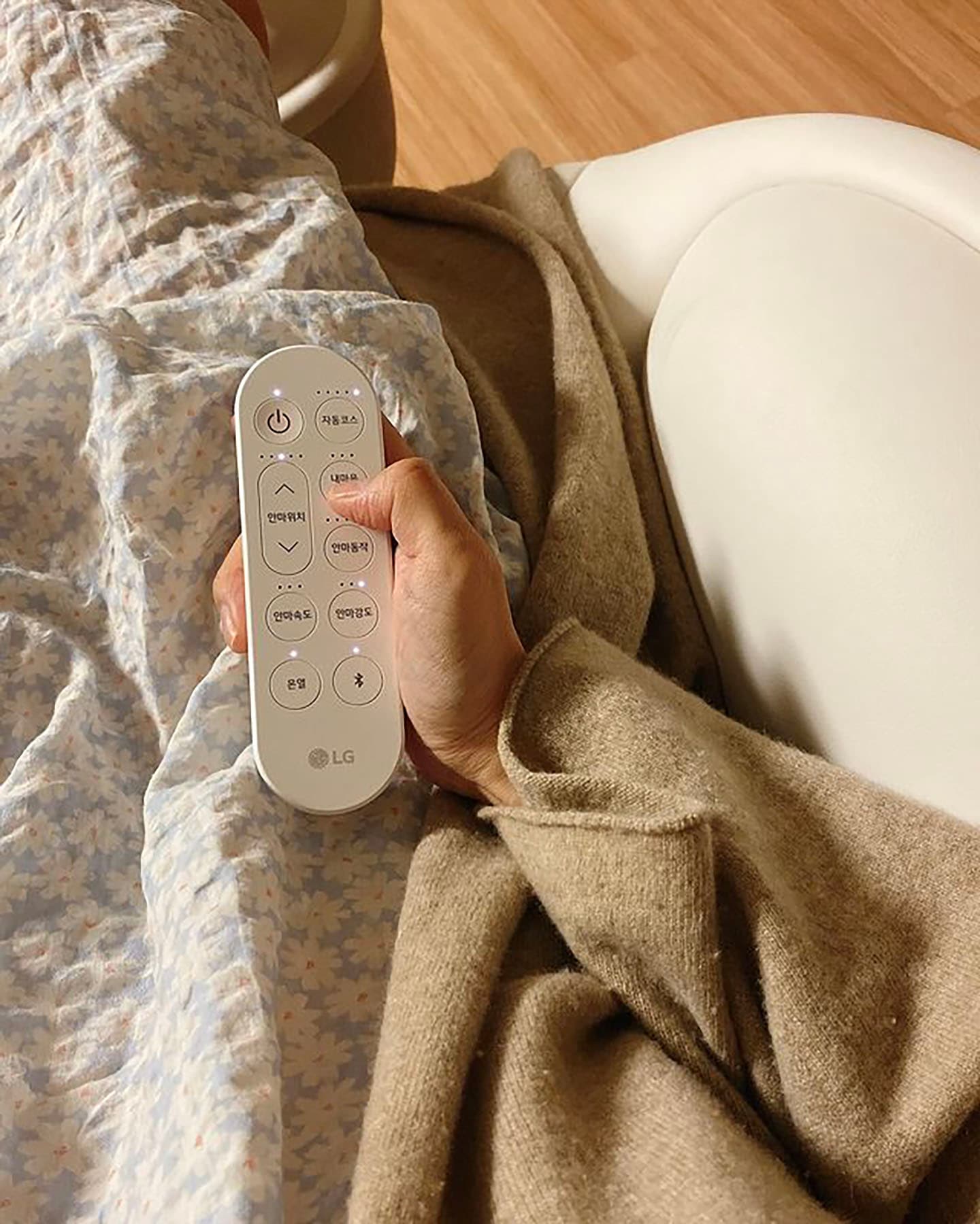 안마 의자에 앉아 포근한 담요를 덮고 무선 리모컨을 조작하는 모습으로, 부드러운 조명 아래 따뜻한 니트 가디건과 플라워 패턴의 이불이 어우러져 아늑한 분위기를 연출하는 이미지