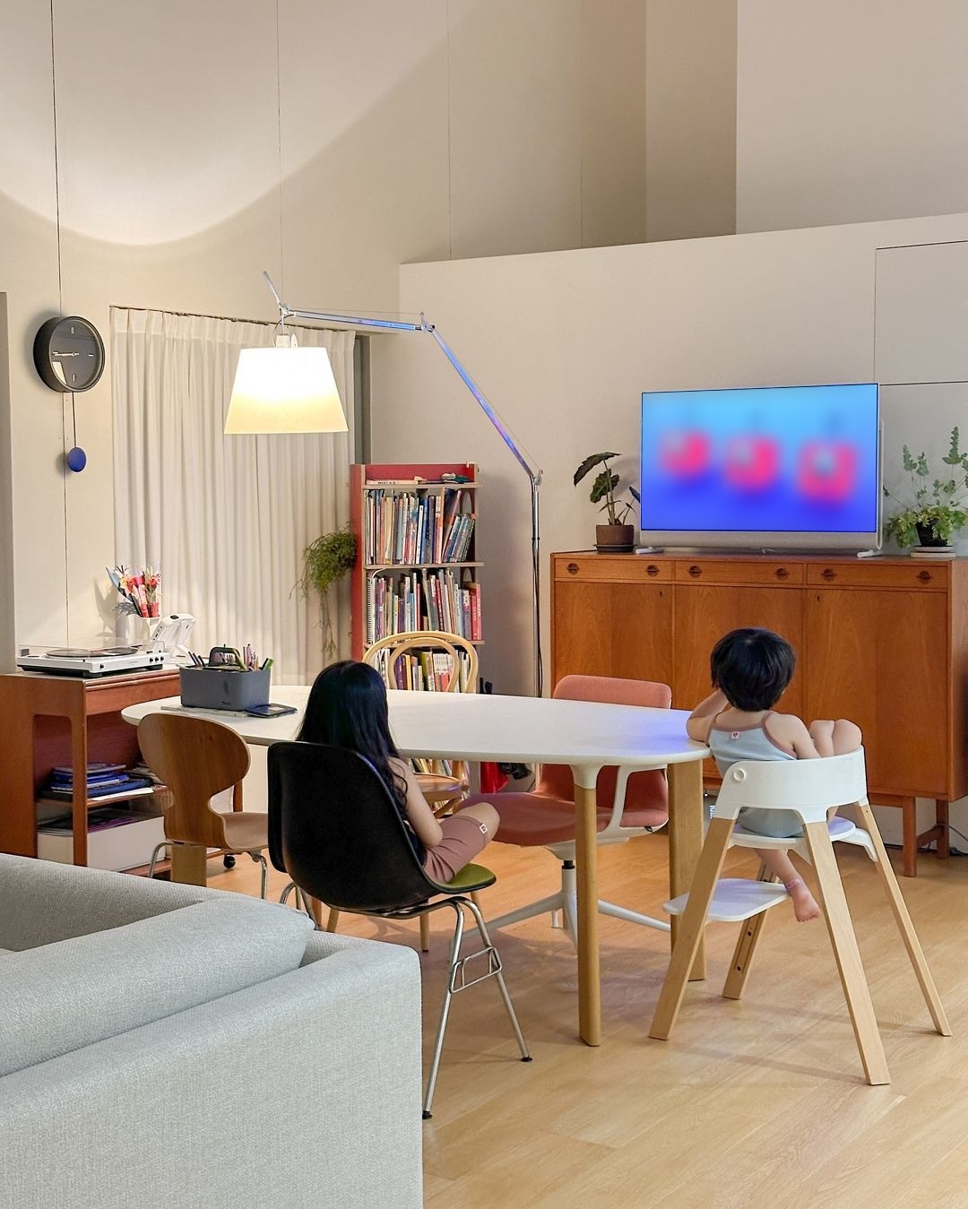 식탁에 앉은 두 아이가 TV를 보고 있음. 주변에는 책장, 스탠드 조명, 장식장 위 화분과 LG 올레드 포제가 놓여 있음. 배경에는 커튼과 시계가 있음.