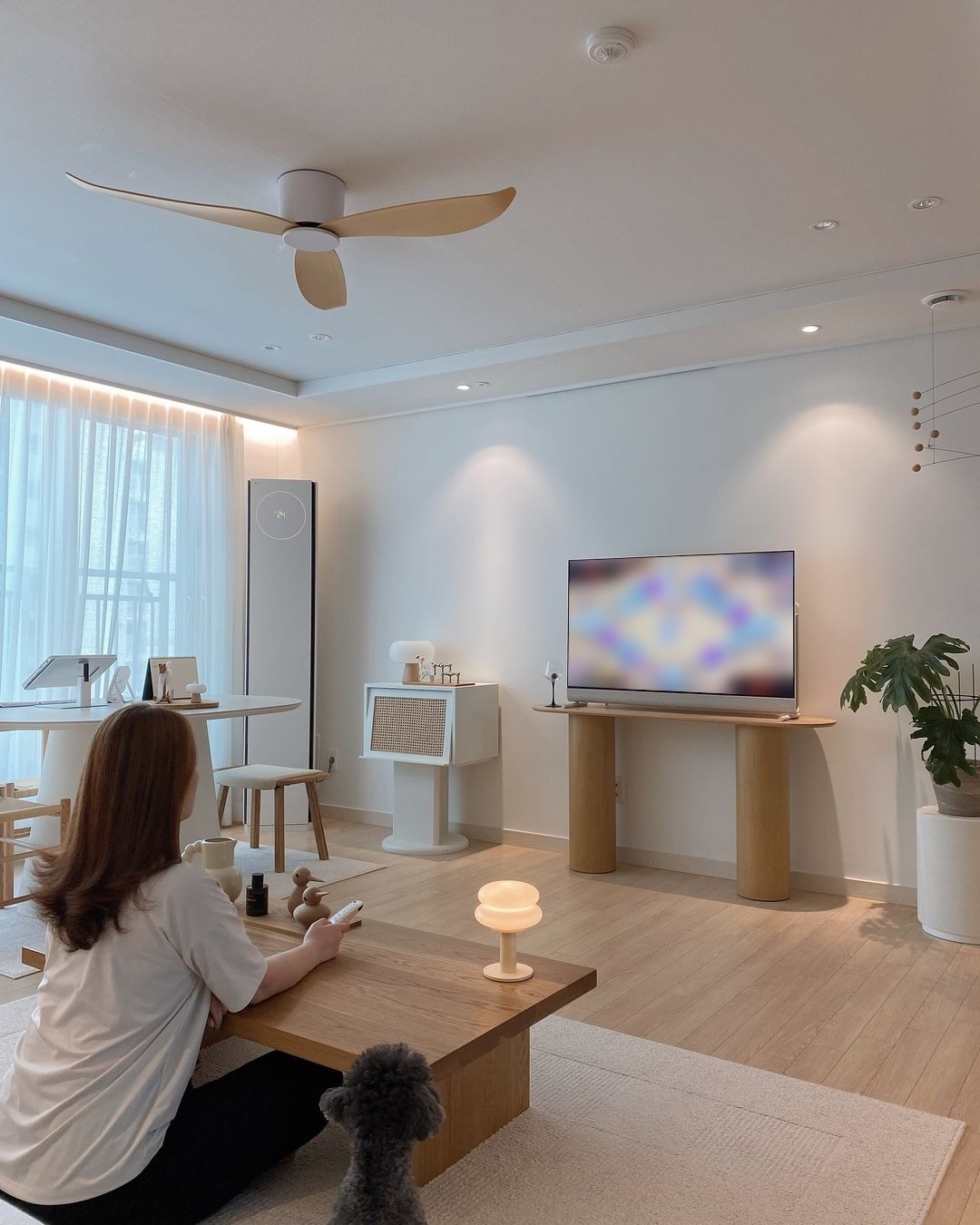 거실에서 한 여성이 무드 등이 있는 테이블에 회색 푸들과 앉아 TV 리모컨을 들고 있음. LG 올레드 포제가 원목 테이블 위에 놓여 있으며, 창가엔 둥근 테이블과 의자, 공기청정기가 있음. 천장엔 나무 날개형 실링팬이 설치됨.