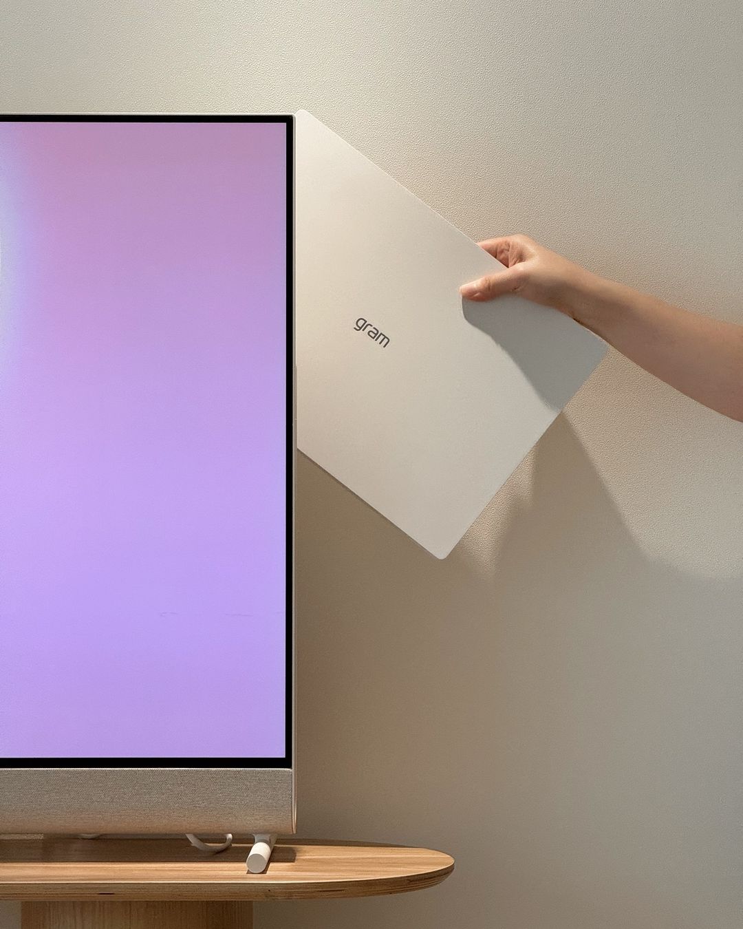 LG 올레드 포제 화면에 연보라색 배경 표시됨. 한 손이 흰색 LG Gram 노트북을 TV 후면 수납 공간에 넣고 있음. 아래에는 원목 테이블과 TV 스탠드 일부 보임.