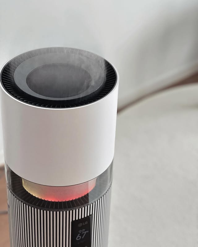 상단에서 수증기가 분사되는 세로 줄무늬 디자인의 퓨리케어 하이드로타워 클로즈업, 디스플레이에 습도(67%)를 표시. 배경에는 밝은 톤의 소파가 있음.