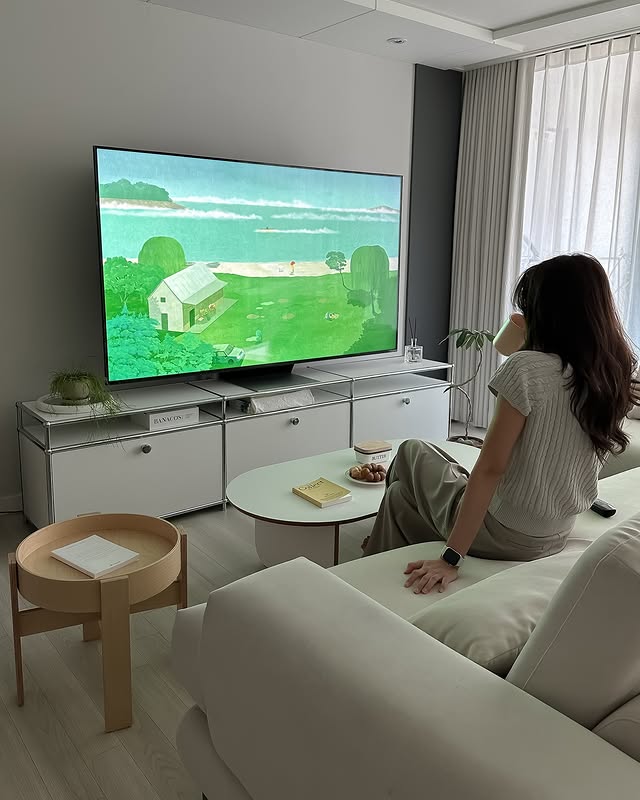 LG QNED evo TV가 거실에 배치되어 있으며, 한 여성이 소파에 앉아 TV 화면을 바라보며 음료를 마시고 있다. TV 화면에는 푸른 자연 풍경과 작은 집이 표시되어 있다.