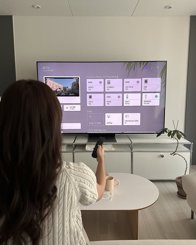 사용자가 LG QNED evo TV를 리모컨으로 조작하는 모습. TV 화면에는 LG ThinQ 홈 대시보드가 표시되어 있으며, 스마트 기능을 제어하는 여러 아이콘이 정렬되어 있다.