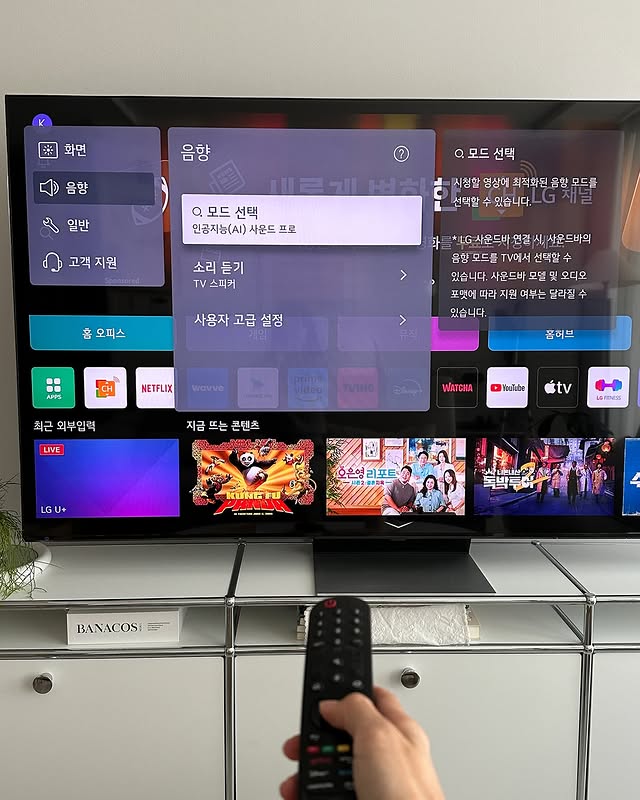 LG QNED evo TV 홈화면에 콘텐츠 서비스가 표시된 모습. 사용자가 리모컨을 들고 화면을 조작하고 있으며, TV 화면에는 인공지능(AI) 사운드 프로 설정 옵션이 활성화되어 있다.
