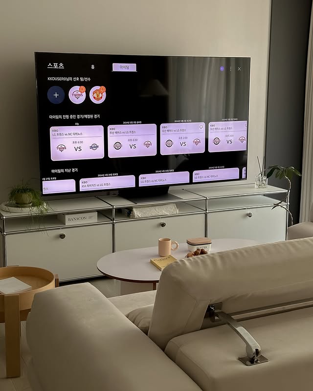 LG QNED evo TV가 거실 공간에 설치되어 있으며, 화면에는 스포츠 경기 일정이 표시되어 있다. TV는 모던한 인테리어와 조화를 이루며, 소파와 커피 테이블이 배치된 편안한 공간을 연출한다.