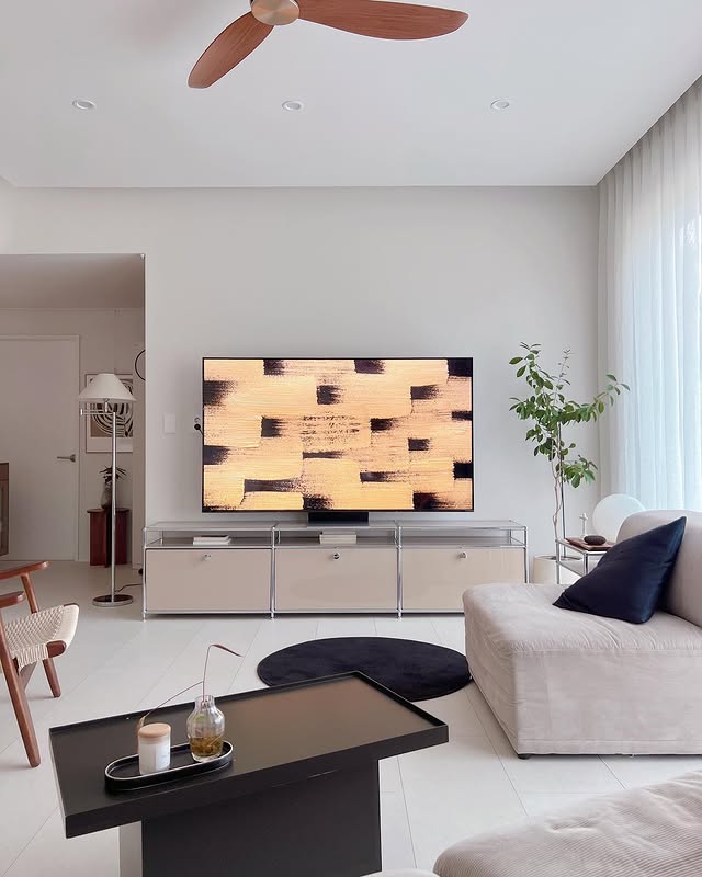 LG QNED evo TV가 거실에 배치된 모습. 벽에 설치된 LG QNED evo TV 화면에는 추상적인 패턴의 예술 작품이 표시되어 있으며, 모던한 인테리어의 거실 공간과 조화를 이루고 있다. TV 아래에는 금속 프레임이 돋보이는 수납장이 배치되어 있으며, 소파와 블랙 원형 러그, 식물이 공간을 아늑하게 연출하고 있다.