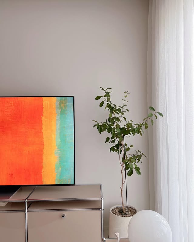 LG QNED evo TV가 거실 한쪽에 배치되어 있으며, 스크린에는 밝은 색감의 추상적인 그래픽이 표시되어 있다. 옆에는 작은 화분이 놓여 있고, 부드러운 커튼이 자연광을 은은하게 투과하는 모습이 보인다.