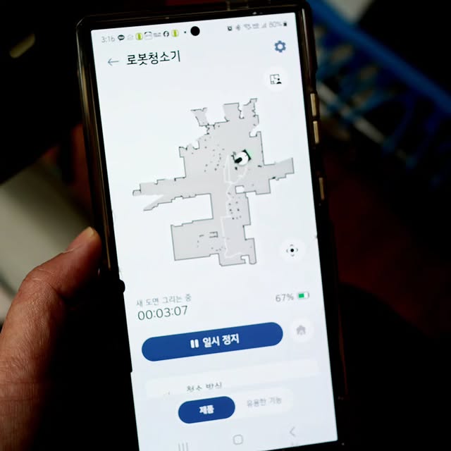 스마트폰 화면에 LG 코드제로 로보킹 AI 올인원의 청소 진행 상태를 실시간으로 확인할 수 있는 ThinQ 앱 화면이 표시된 모습. 손으로 스마트폰을 잡고 있으며, 지도 기반의 청소 영역이 표시됨.