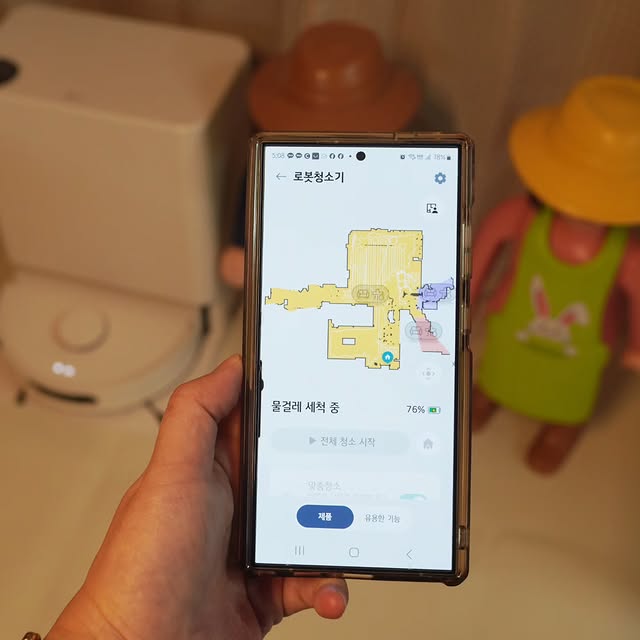 스마트폰 화면에 표시된 LG 코드제로 로보킹 AI 올인원의 청소 맵. 물걸레 세척 모드가 실행 중이며, 배경에는 로봇청소기와 도킹 스테이션, 장난감 인형이 보임.