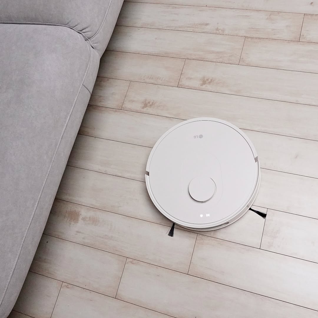 LG 코드제로 로보킹 AI 올인원이 밝은 나무 바닥에서 소파 근처를 청소하는 모습. 원형의 흰색 본체에 레이저 센서가 탑재되어 있으며, 바닥에 있는 먼지를 정밀하게 감지하며 이동하는 장면이다.