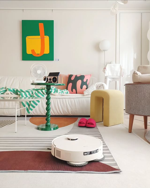 LG 코드제로 로보킹 AI 올인원이 거실 러그 위를 청소하는 모습. 감각적인 인테리어의 밝은 거실에 소파, 컬러풀한 쿠션, 디자인 테이블, 초록색 그림 등이 조화를 이루고 있다. 