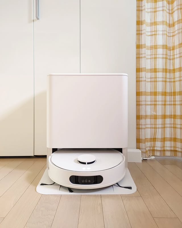 LG 코드제로 로보킹 AI 올인원이 전용 충전 도킹 스테이션에 장착된 모습. 깔끔한 흰색 디자인의 청소기가 먼지 자동 비움 기능을 수행하는 중이며, 배경에는 미니멀한 옷장과 노란 체크 패턴의 커튼이 보인다. 