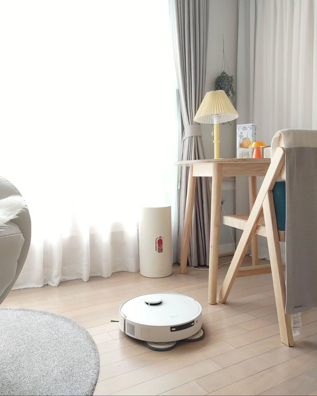 LG 코드제로 로보킹 AI 올인원이 햇살이 들어오는 창가 근처에서 바닥을 청소하는 모습. 우드 톤 가구와 크림색 커튼이 조화롭게 배치된 아늑한 공간에서 로봇청소기가 부드럽게 이동하며 물걸레 청소를 진행하고 있다.
