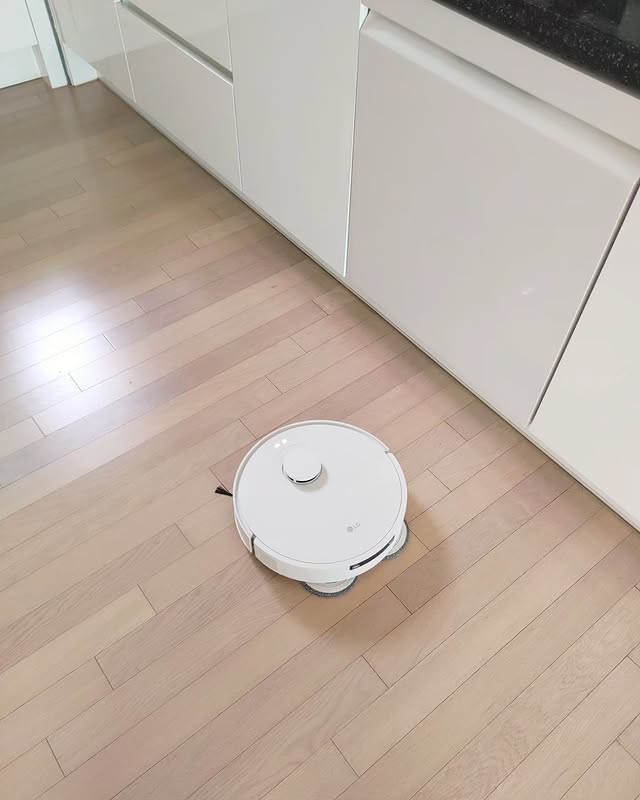 LG 코드제로 로보킹 AI 올인원이 화이트 톤 주방 바닥을 청소하는 모습. 빌트인 수납장이 깔끔하게 정돈된 공간에서 로봇청소기가 자동으로 이동하며 물걸레 청소를 수행하고 있어 위생적인 주방 환경을 강조하는 장면.