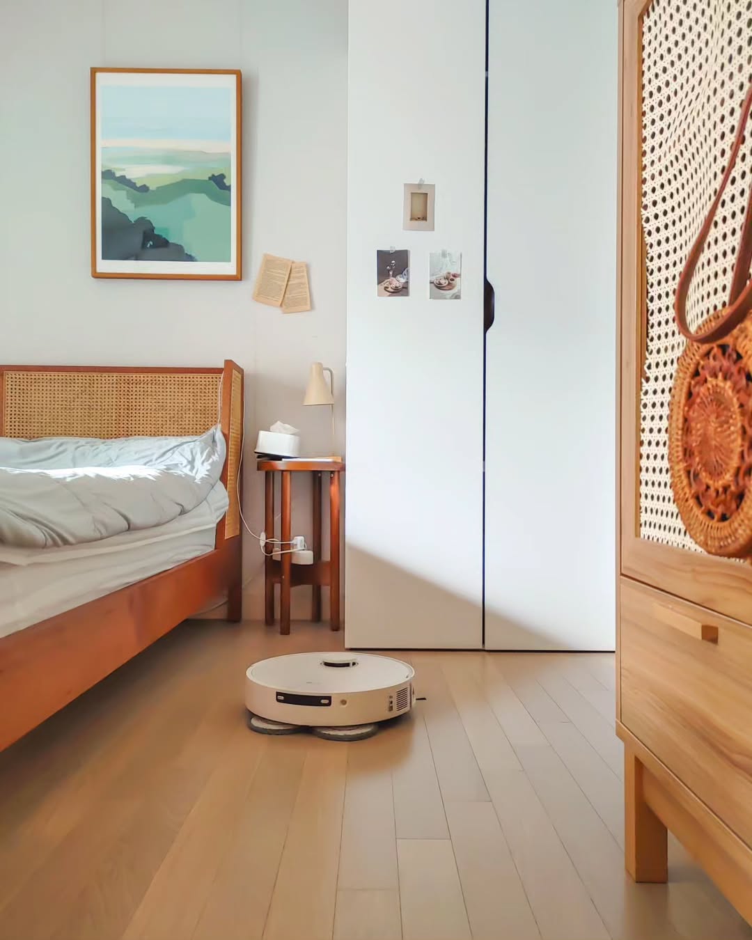 LG 코드제로 로보킹 AI 올인원이 우드톤 가구로 꾸며진 침실에서 바닥을 청소하는 모습. 침대 옆에는 작은 협탁과 스탠드 조명이 놓여 있으며, 벽에는 자연 풍경이 그려진 액자가 걸려 있다.