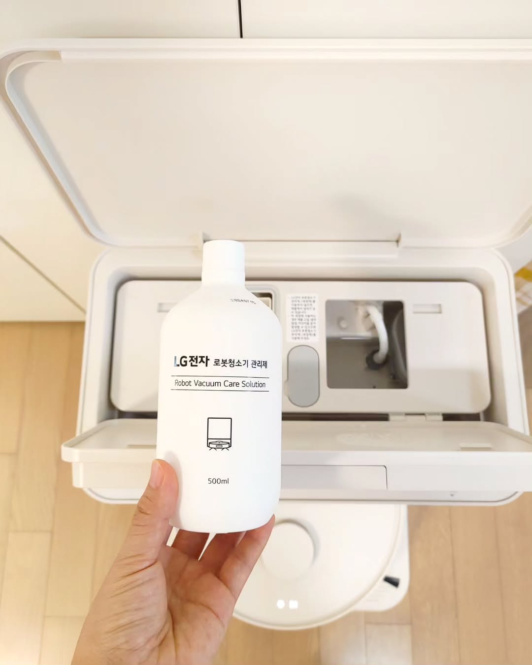 LG 코드제로 로보킹 AI 올인원의 관리 액세서리인 LG전자 로봇청소기 관리제 500ml 용기를 손으로 들고 있는 모습. 배경에는 로봇청소기의 충전 도킹 스테이션 내부가 보이며, 청소기를 효율적으로 유지·관리할 수 있는 솔루션을 강조하는 장면.