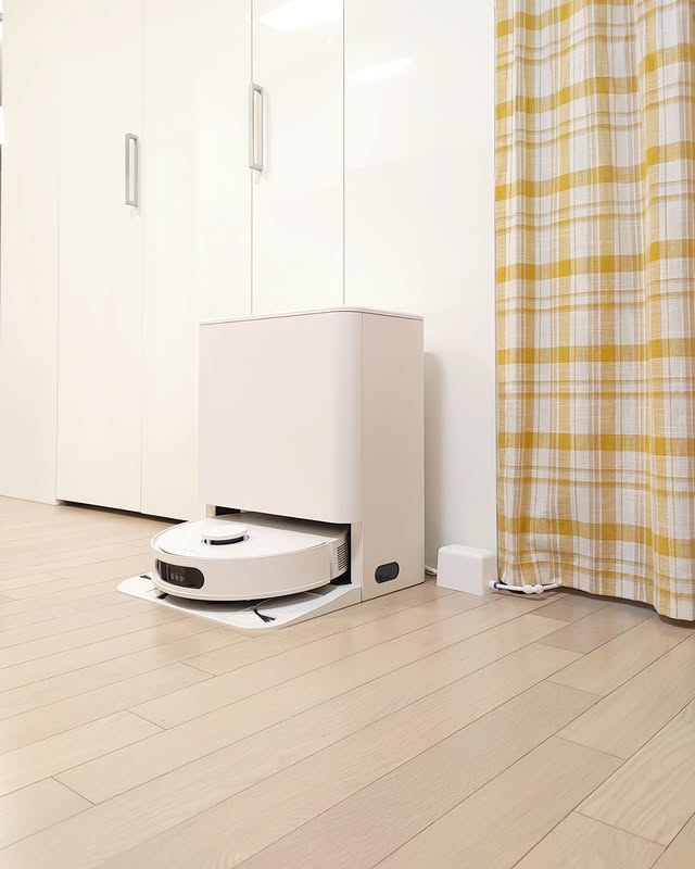 LG 코드제로 로보킹 AI 올인원이 전용 충전 도킹 스테이션에서 충전 중인 모습. 깔끔한 화이트톤의 공간에 노란 체크 패턴의 커튼이 조화를 이루며, 로봇청소기가 스마트하게 관리되는 모습을 보여준다.