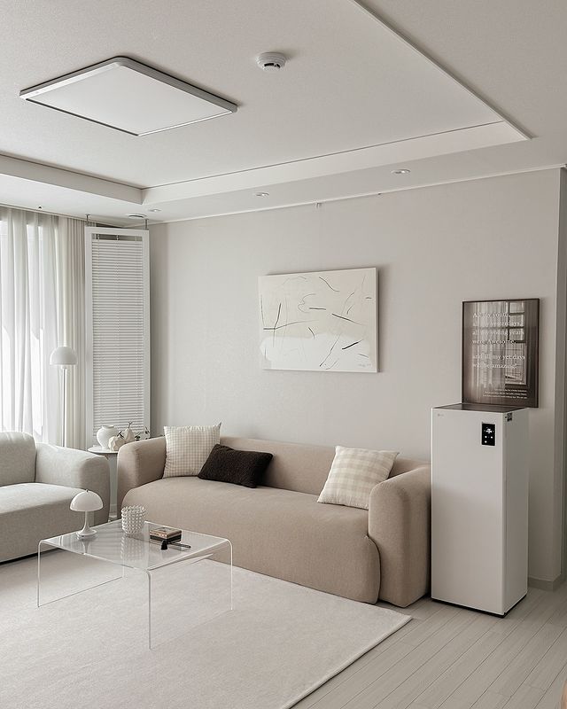 베이지 톤 소파와 투명한 유리 테이블이 조화를 이루는 화이트톤 거실에 스타일러 오브제컬렉션 슈케어가 배치되어 있음. 소파 위에는 베이지와 검정 쿠션이 놓여 있고, 벽에는 그림들이 장식됨.