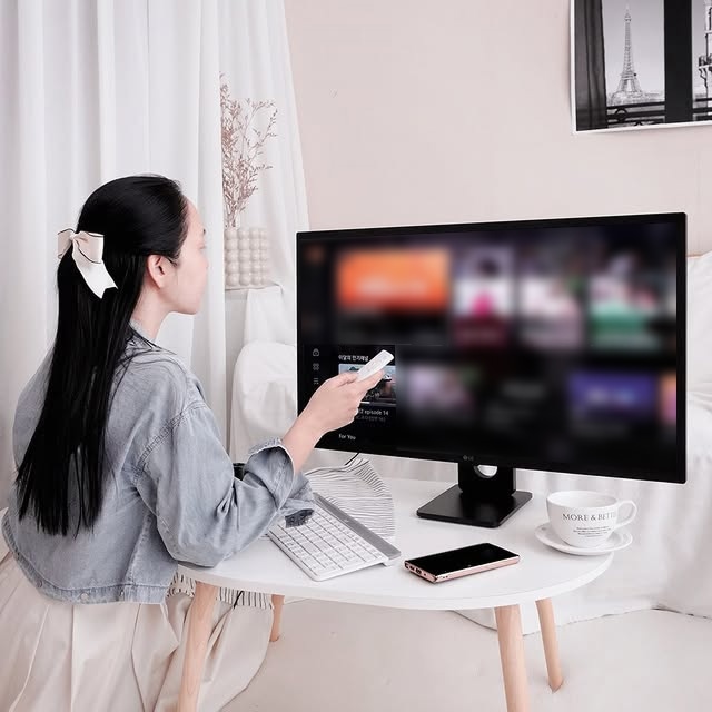 한 여성이 LG 스마트모니터 앞에 앉아 리모컨으로 화면을 조작하며 콘텐츠를 감상하고 있다. 책상이 놓여 있는 공간은 따뜻한 분위기의 인테리어로 꾸며져 있으며, 흰색 커튼과 액자가 배경에 보인다.