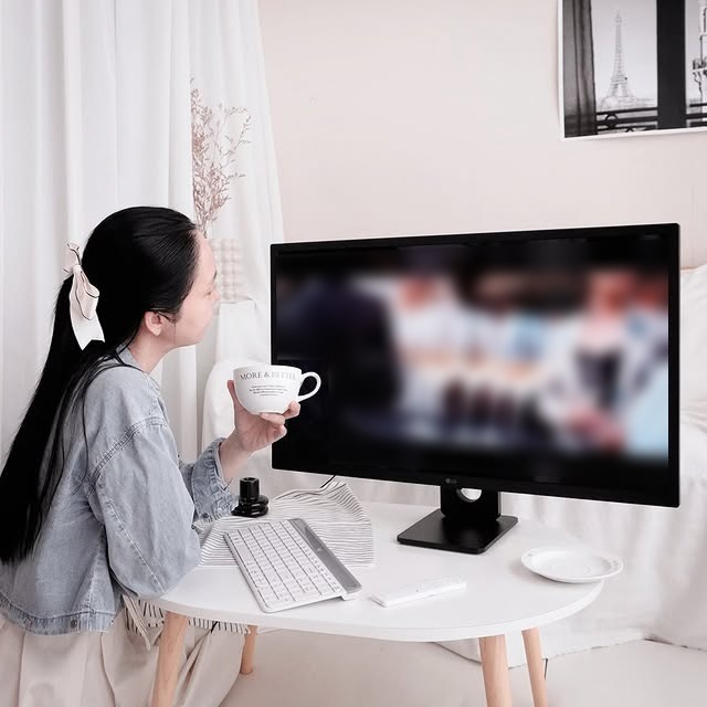 여성이 LG 스마트모니터 화면을 보며 커피를 들고 여유롭게 감상하고 있다. 화면에는 영상이 재생 중이며, 책상 위에는 키보드와 리모컨이 놓여 있다. 배경에는 흰색 커튼과 벽에 걸린 액자가 보인다.