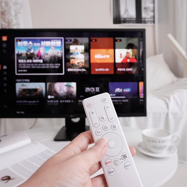 사용자의 손이 LG 스마트모니터 전용 매직 리모컨을 들고 있으며, 모니터 화면에는 넷플릭스, 디즈니 플러스, 유튜브 등의 앱 아이콘과 다양한 추천 콘텐츠가 표시되어 있다. 책상 위에는 문서와 커피잔이 놓여 있으며, 배경에는 따뜻한 톤의 실내 인테리어가 보인다.