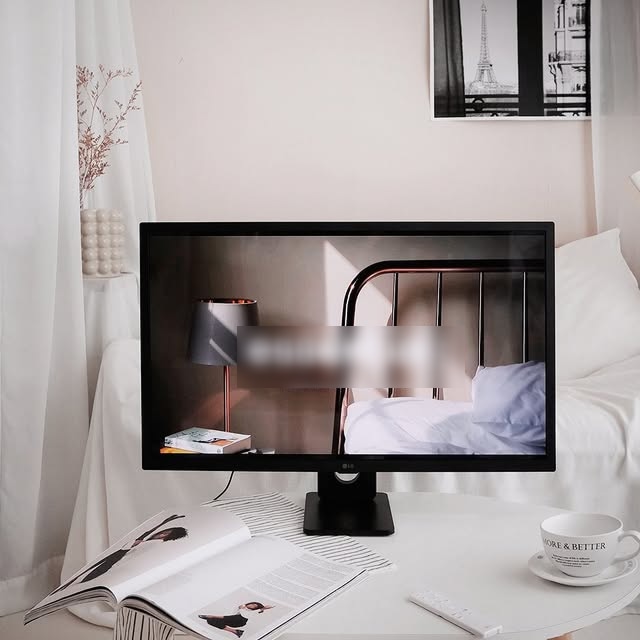 LG 스마트모니터가 책상 위에 놓여 있으며, 화면에는 모던한 침실 장면이 표시되어 있다. 책상이 있는 공간은 아늑한 인테리어로 꾸며져 있으며, 커피잔과 잡지가 함께 놓여 있어 편안한 분위기를 연출한다.