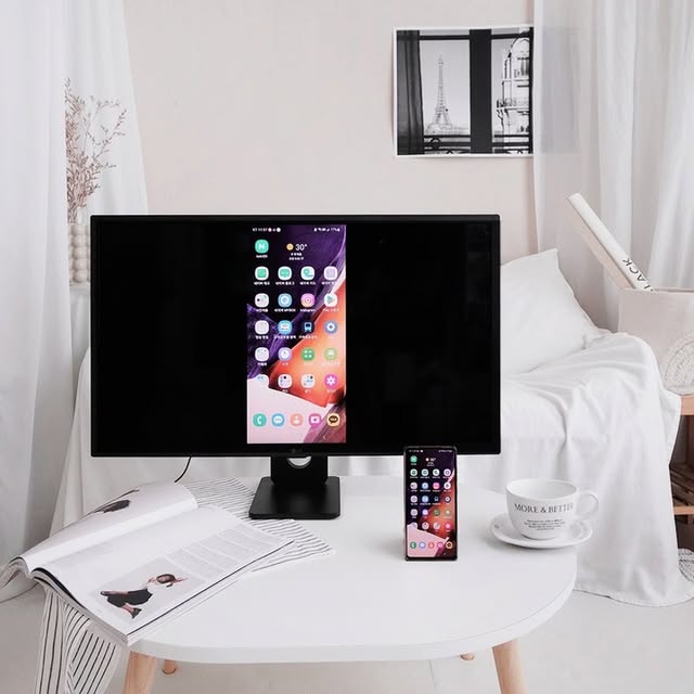 LG 스마트모니터가 책상 위에 놓여 있으며, 스마트폰과 무선으로 연결되어 화면 미러링 기능이 실행된 모습이다. 스마트폰 화면이 모니터에 그대로 표시되며, 깔끔한 인테리어 속에서 무선 연결의 편리함을 강조하는 장면이다.