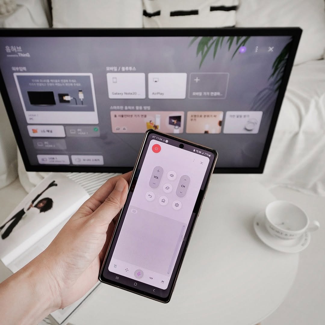 LG 스마트모니터가 책상 위에 놓여 있으며, 사용자가 스마트폰을 활용해 원격으로 모니터를 제어하는 모습이다. 스마트폰 화면에는 리모컨 인터페이스가 표시되어 있고, 모니터 화면에는 다양한 설정 옵션이 보이며, 스마트 기능을 활용한 간편한 조작이 강조되고 있다.