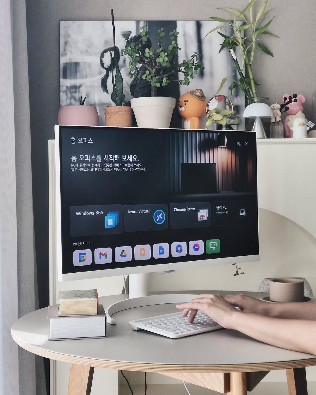 LG 스마트 모니터가 책상 위에 배치되어 있으며, 화면에는 ‘홈 오피스’ 기능이 실행된 모습이 보인다. 사용자가 키보드를 이용해 원격 PC 기능을 설정하고 있으며, 모니터 뒤쪽에는 여러 개의 화분과 장식품이 놓여 있다.
