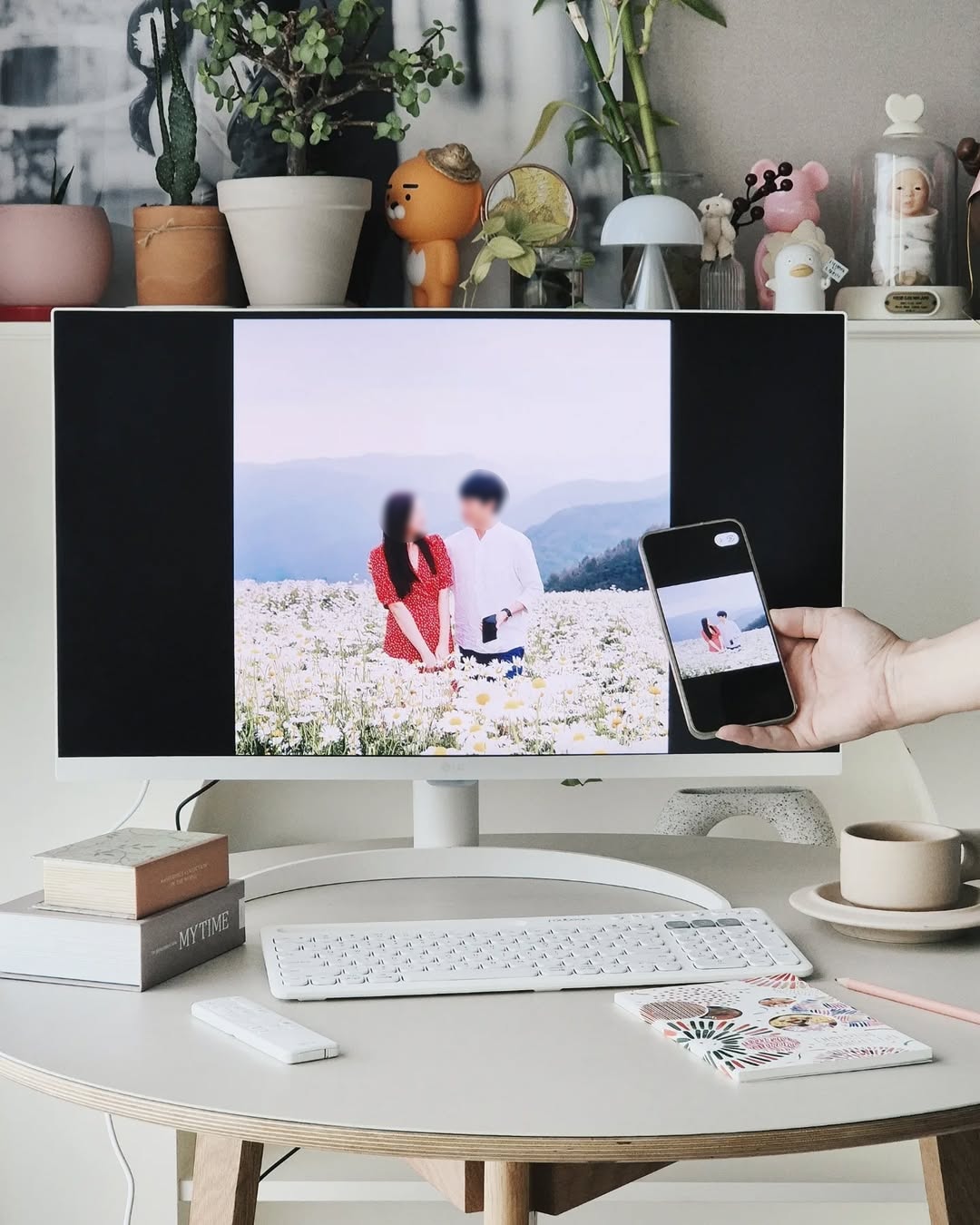  LG 스마트 모니터에서 스마트폰과 화면 미러링 기능을 활용하는 장면. 사용자가 스마트폰으로 찍은 꽃밭에서 연인이 함께 있는 사진을 모니터로 전송하여 감상하는 모습. 책상 위에는 키보드, 리모컨, 노트, 연필, 커피잔 등이 함께 놓여 있다.