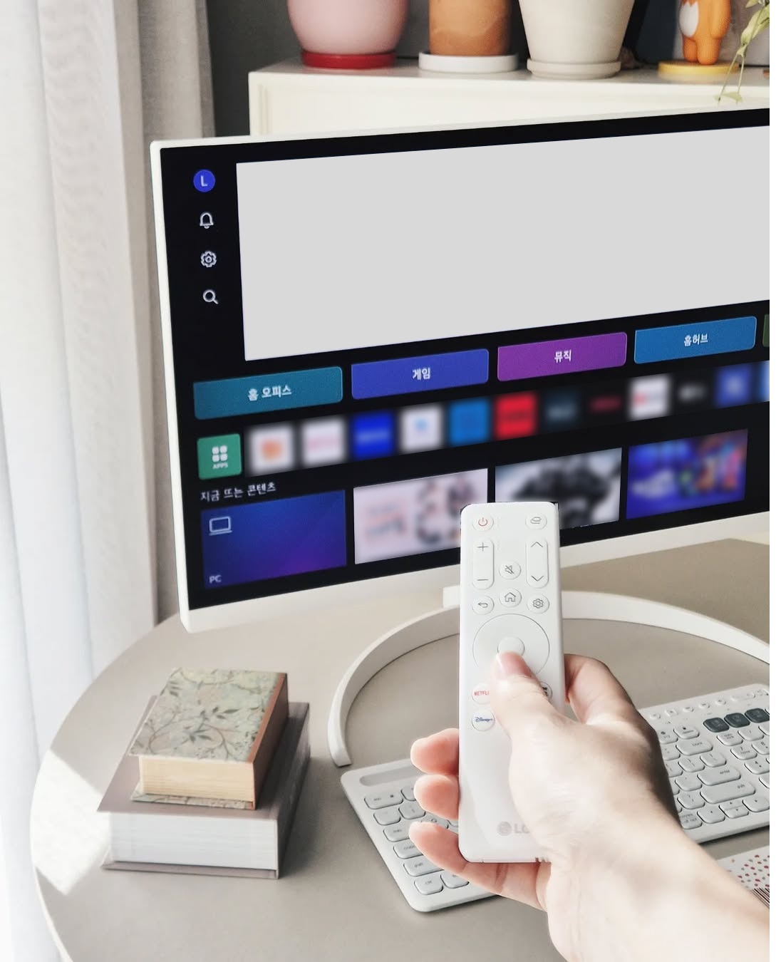 LG 스마트 모니터가 책상 위에 놓여 있으며, 사용자가 손으로 리모컨을 조작하고 있다. 모니터 화면에는 다양한 콘텐츠 아이콘과 메뉴들이 표시되어 있어 스마트 기능을 활용하는 장면을 보여준다.