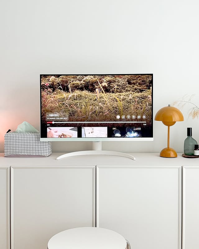 LG 스마트 모니터가 거실의 깔끔한 인테리어 속에 배치되어 있으며, 화면에는 자연 풍경과 동영상 스트리밍 서비스 UI가 표시되고 있다. 모니터 아래에는 곡선형 스탠드가 있으며, 오른쪽에는 노란색 스탠드 조명과 인테리어 소품들이 놓여 있다.