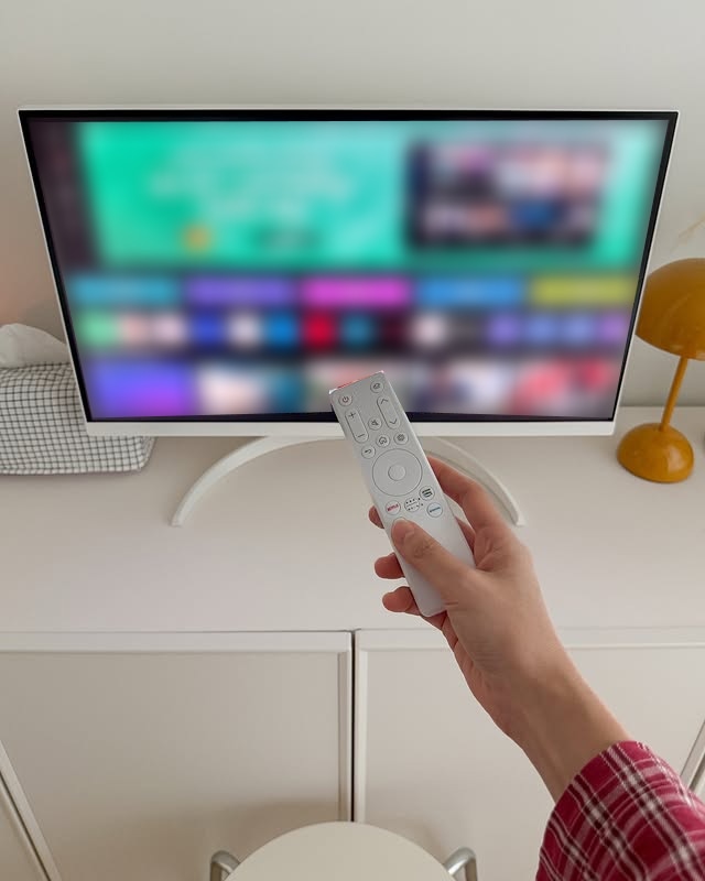 한 손이 LG 스마트 모니터의 리모컨을 잡고 화면을 조작하고 있다. 모니터는 화이트 색상의 곡선형 스탠드 위에 올려져 있으며, 화면에는 스마트 TV 인터페이스가 보인다. 오른쪽에는 노란색 스탠드 조명이 배치되어 있다.