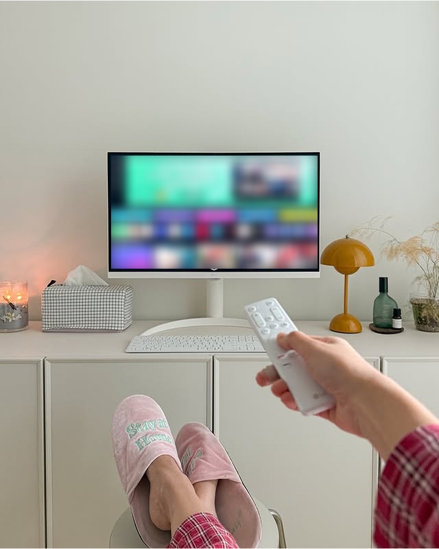 사용자가 핑크색 실내화를 신고 다리를 올린 채 LG 스마트 모니터를 리모컨으로 조작하며 콘텐츠를 감상하는 모습. 모니터 주변에는 감성적인 인테리어 소품들이 배치되어 있다.