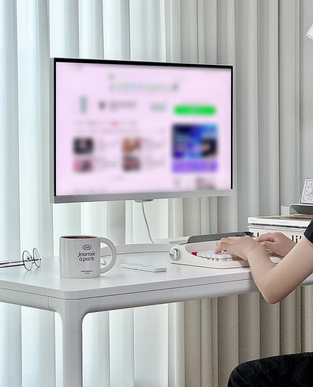 흰색 책상에 놓인 LG 스마트 모니터에서 웹사이트가 열려 있고, 사용자가 키보드를 사용해 작업을 하고 있다. 창가의 흰색 커튼이 자연광을 비추고 있으며, 책상 위에는 머그컵과 리모컨이 놓여 있다.