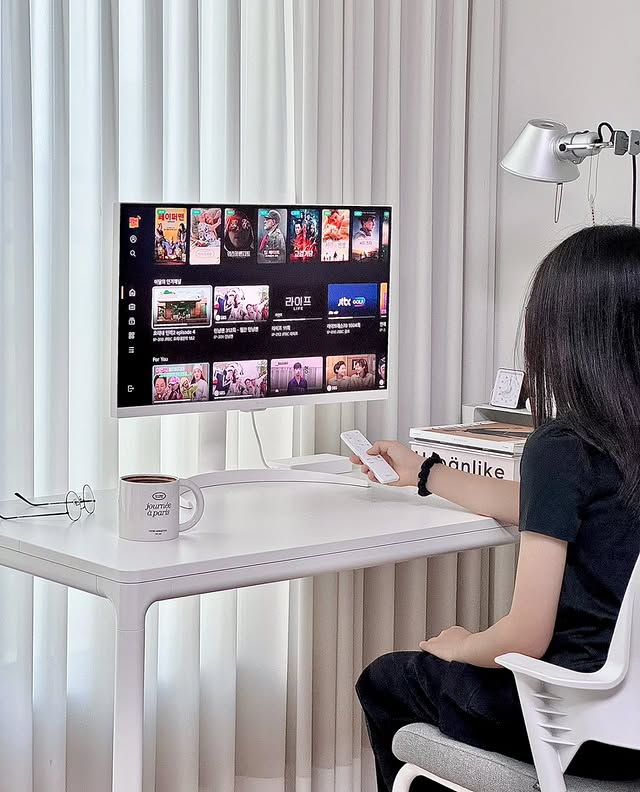 사용자가 흰색 책상에 앉아 LG 스마트 모니터로 OTT 콘텐츠를 시청하고 있다. 검은색 상의의 사용자가 리모컨을 들고 있으며, 커피잔과 책이 책상 위에 정리되어 있다.