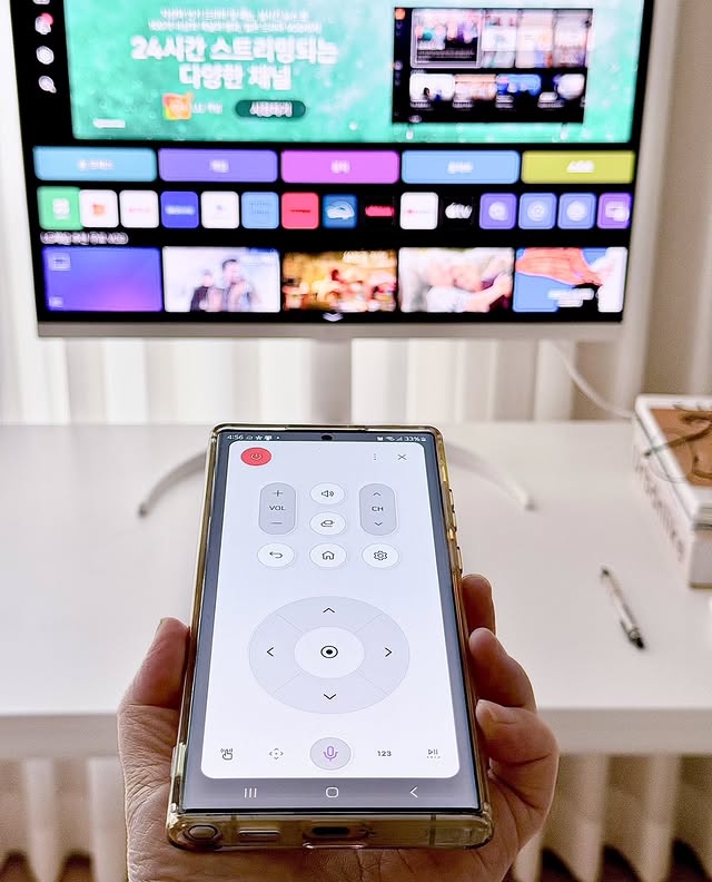스마트폰 화면에 LG 스마트 모니터의 원격 조정 할 수 있는 ThinQ 앱이 실행된 모습. 사용자의 손이 스마트폰을 들고 있으며, 배경에는 모니터 화면에 OTT 콘텐츠 목록이 표시되고 있다.