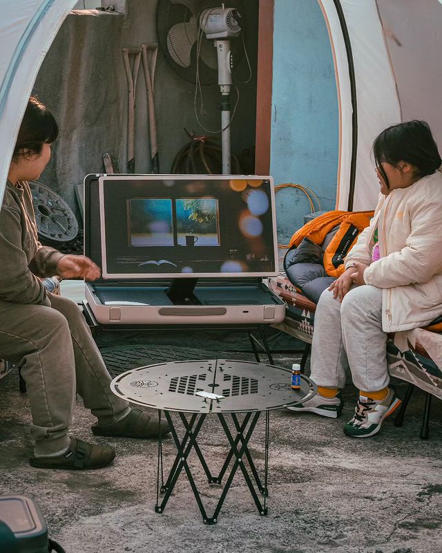 어린이 두 명이 캠핑 텐트 안에서 스탠바이미 Go 화면을 보며 앉아 있음. 화면에는 창문과 컵이 있는 풍경이 보임. 테이블 위에는 음료수 병과 캠핑용 접이식 테이블이 있음.