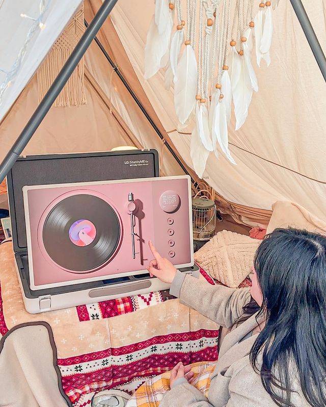 캠핑 텐트 안에서 한 사람이 스탠바이미 Go 화면을 터치하고 있음. 화면에는 턴테이블 인터페이스 표시됨. 주변에 침구류와 장식품 있음.