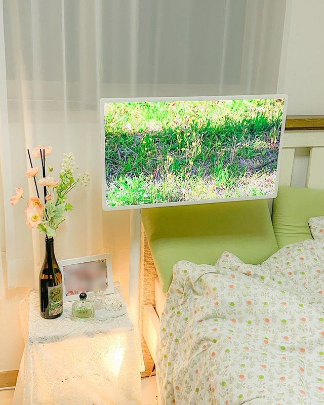 침대 옆 스탠바이미에 초록색 풀밭 장면 표시됨. 옆에는 꽃병과 조명이 있는 협탁 있음. 침대에는 녹색 쿠션과 꽃무늬 이불 있음.