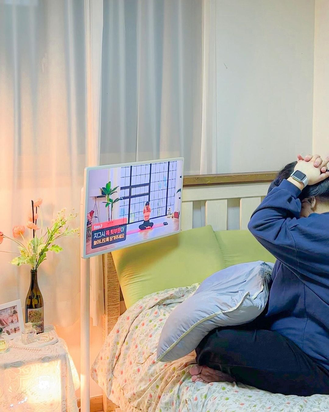 침대 옆 스탠바이미에 요가 강의 화면 표시됨. 한 사람이 침대에 앉아 따라 하고 있음. 옆에는 꽃병과 조명이 있는 협탁 있음. 침대에는 녹색 쿠션과 꽃무늬 이불 있음.