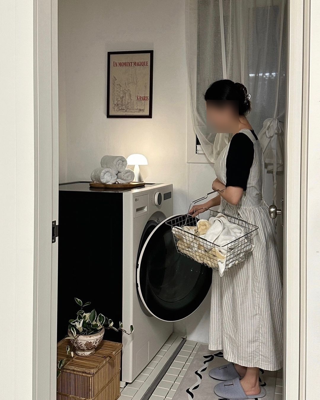 문이 열린 세탁실에 워시콤보 앞에서 바구니에 세탁물을 담고 있는 여성이 문을 열고 있음. 상단에는 수건들과 켜진 무드등이 배치, 벽엔 파리 감성 액자가 있음. 바닥엔 러그와 라탄 박스 위 화분이 놓여 있음.