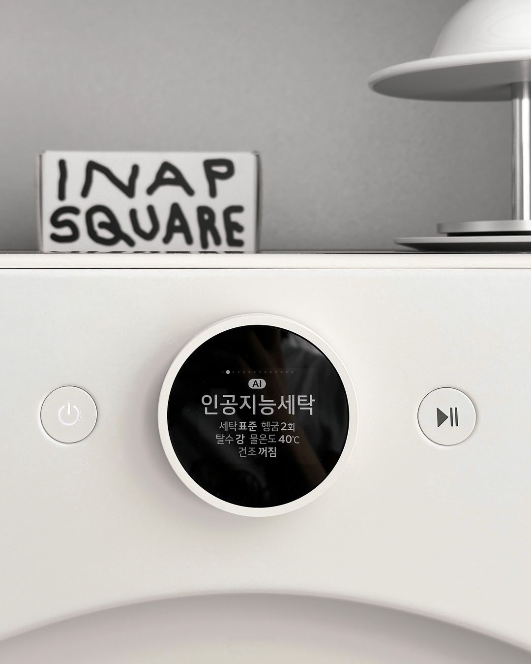 워시콤보의 인공지능세탁 모드와 세부 설정 화면이 표시되어 있고 세탁기 위에는 작은 장식품과 램프가 있음.