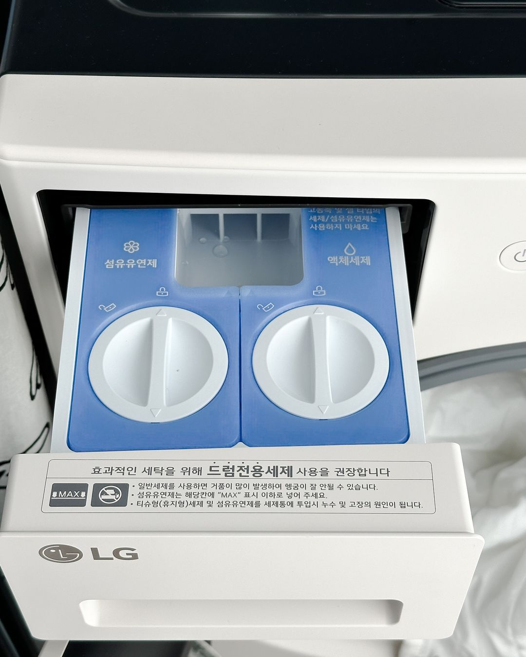 워시콤보의 세제 투입구가 열린 모습으로, 섬유유연제와 액체세제 칸이 분리되어 있음. 각 칸에는 용량 조절 다이얼이 있으며, 아래에는 드럼 전용 세제 사용을 권장하는 안내 문구가 부착됨.
