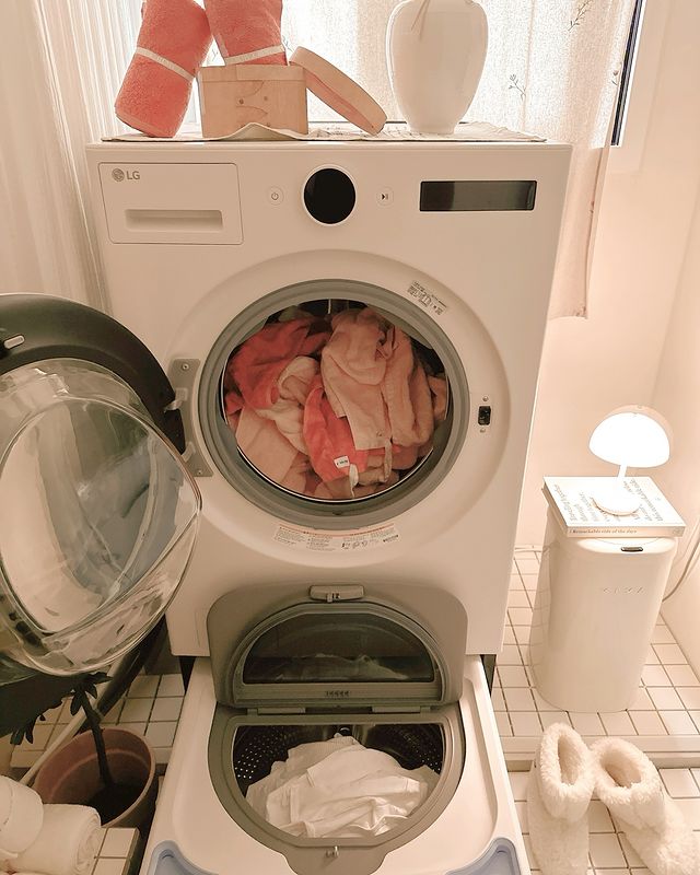 도어가 열린 워시타워와 워시타워 미니의 내부 모습으로, 상단 드럼에는 오렌지색 타월이, 하단 드럼에는 흰색 의류가 들어 있음. 배경에는 레이스 커튼이 있는 창과 켜진 무드등, 털부츠가 놓여 아늑한 분위기를 연출함.